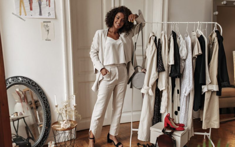 Full-length portrait of stylish dark-skinned curly woman in white pants, jacket and top posing in dressing room. Young lady leans on hanger.